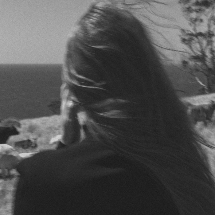 A person with long hair stands with their back to the camera, enjoying the ocean breeze and serene coastal views on a sunny day near the cliffs edge.